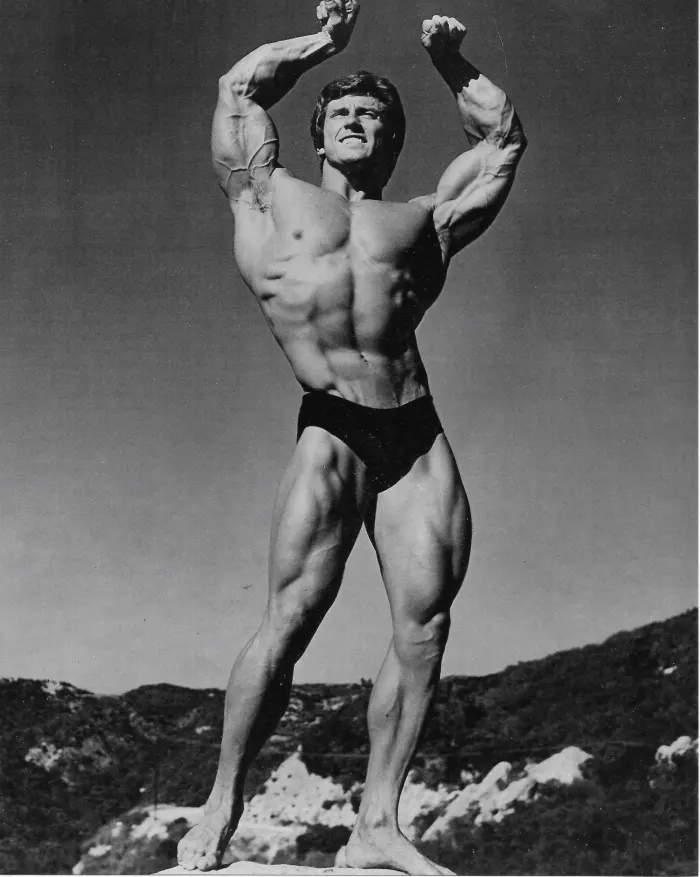 A young Frank Zane raises his arms atop a Malibu rock outcrop, a dramatic black-and-white silhouette against the open sky.
