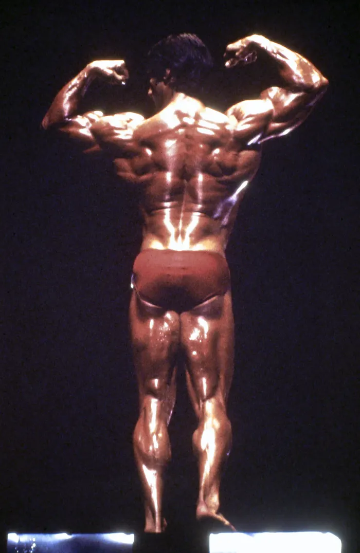 A rear double-biceps pose under real competition-stage spotlights showcases the back development that defined his physique.