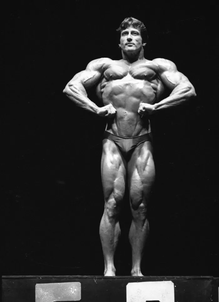 A classic front most-muscular stage pose, direct gaze, hands at the waist.