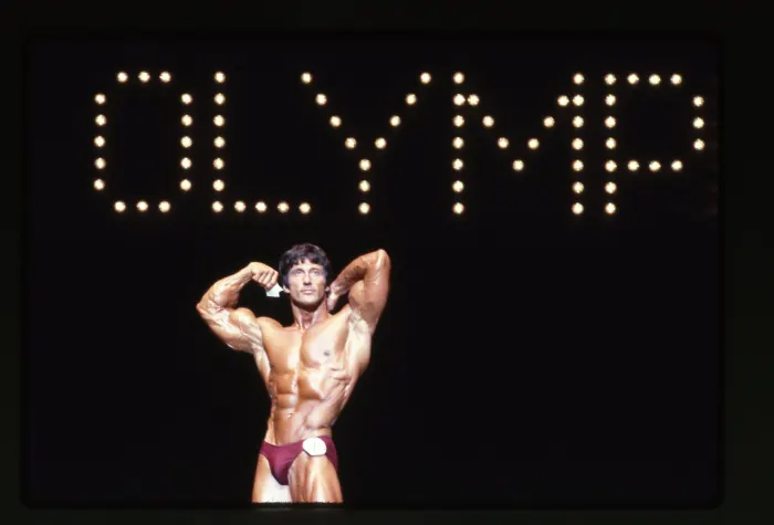 A solo biceps pose directly beneath the illuminated 'OLYMPIA' marquee, unmistakably iconic.