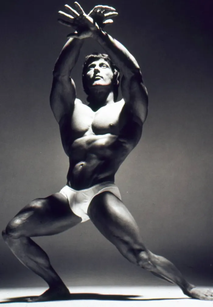 Crossed overhead arms and sculptural lighting recall fine-art studio photography at its most striking.