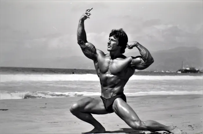 A dynamic side-lunge double-biceps pose on the beach with ocean waves and a distant boat.