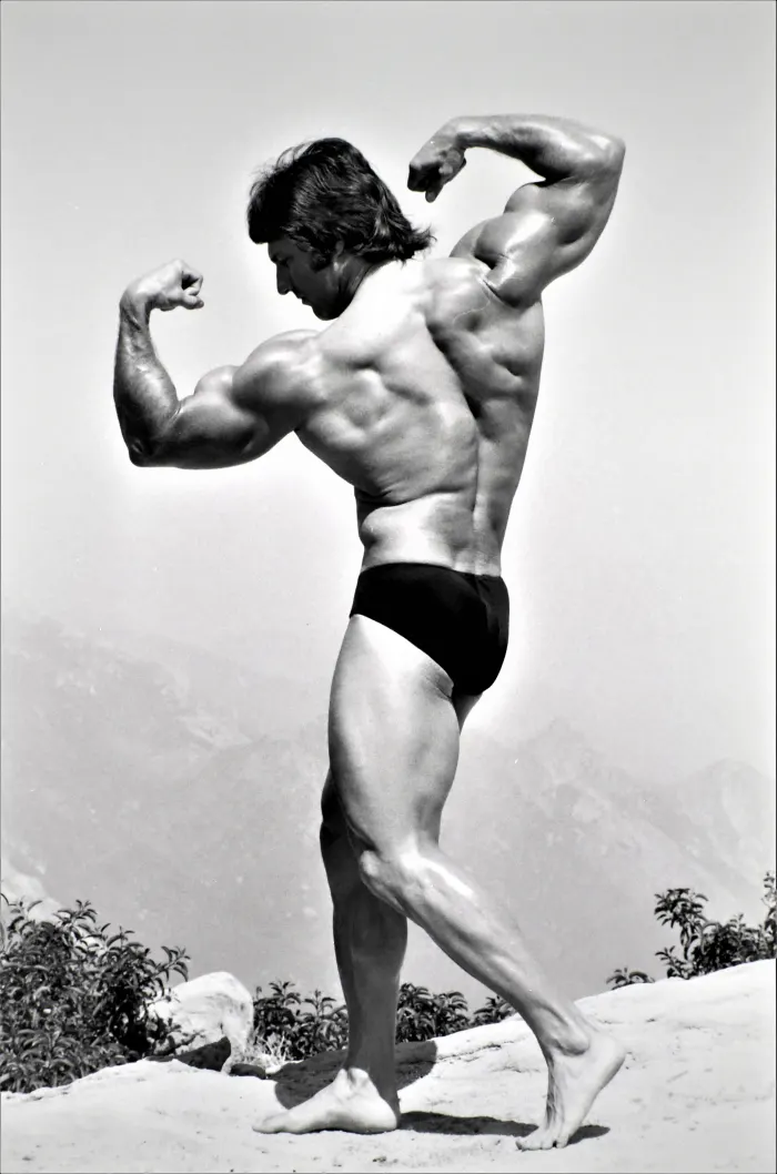 A rear double-biceps pose in the desert, arms fully extended and fingers splayed against open sky.