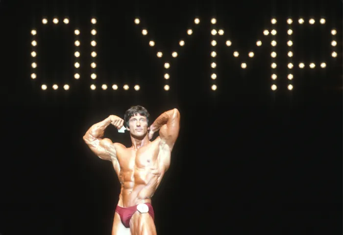 Posing beneath the illuminated 'OLYMPIA' signage, competitor number visible, unmistakably tied to the Mr. Olympia stage.