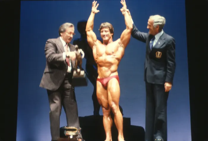 Arms raised as an official presents a trophy and another celebrates beside him, a genuine victory narrative caught mid-moment.