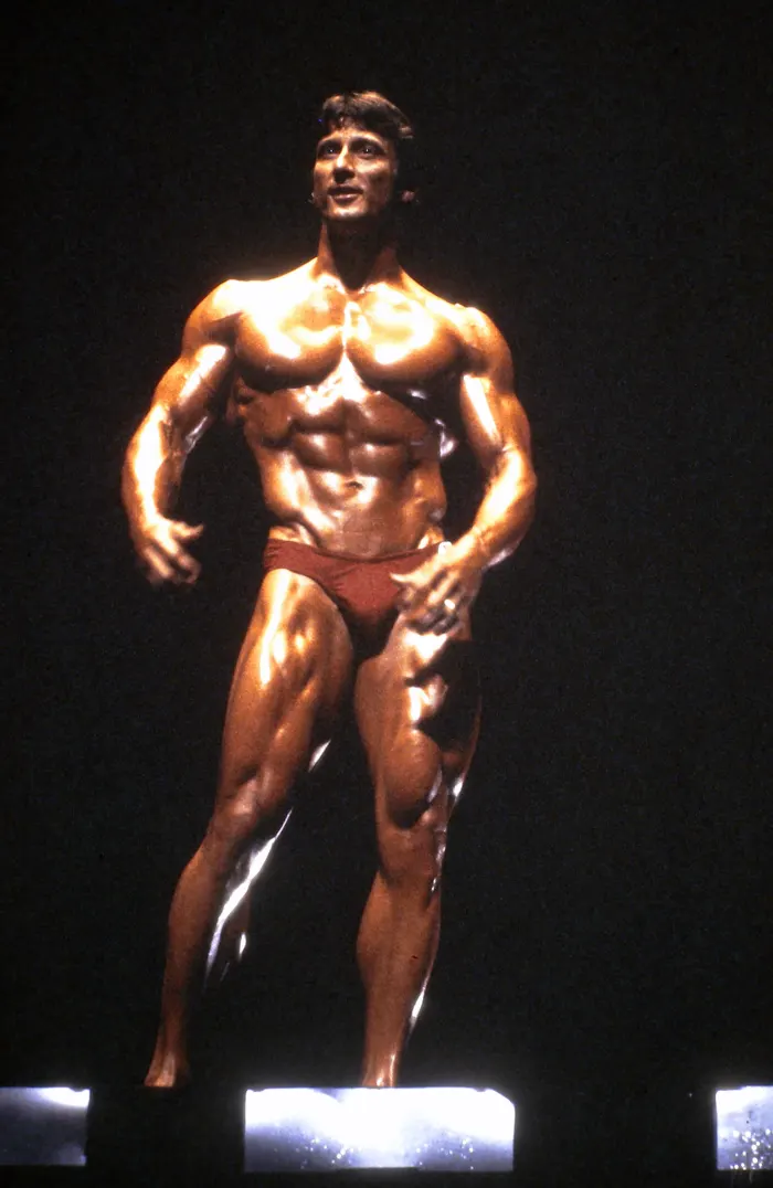 A front-and-center Olympia stage shot with the winner's trophy statue visible in the foreground.