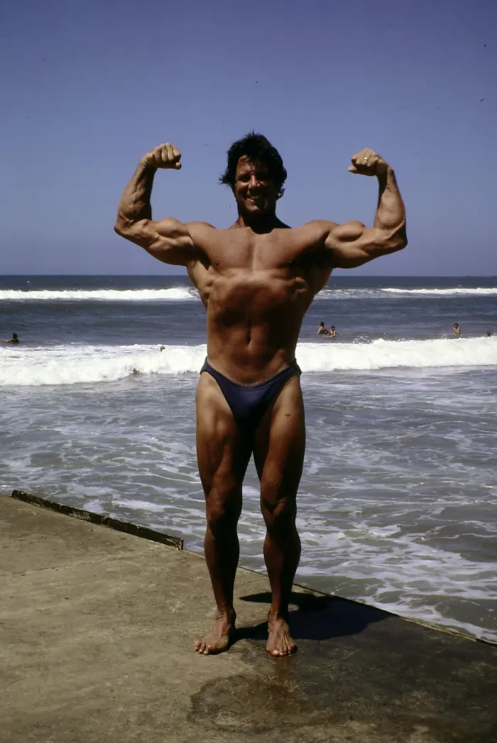 A front double-biceps pose on the beach with a wide, genuine smile and dramatic ocean and sky backdrop.