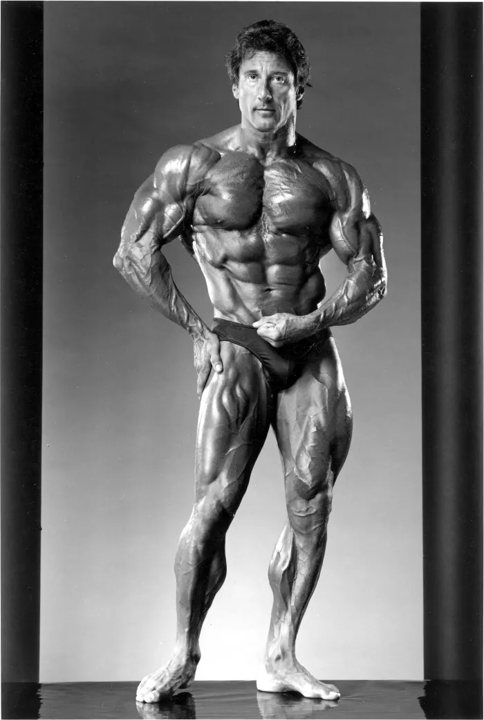 An iconic studio black-backdrop physique pose under theatrical lighting.