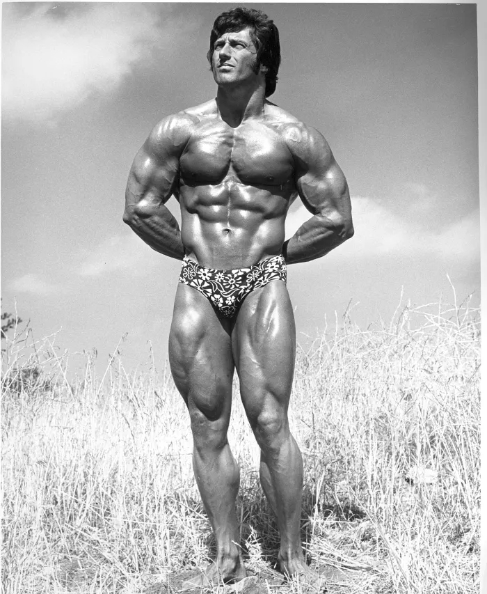 A black-and-white outdoor shot in windswept dry grass under a dramatic sky, iconic floral posing trunks.
