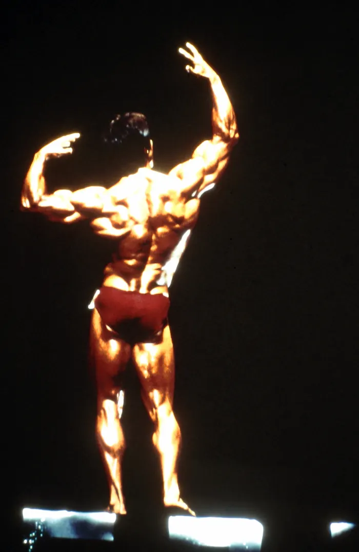 A dramatic rear double-biceps silhouette under hot stage lighting against black backdrop.