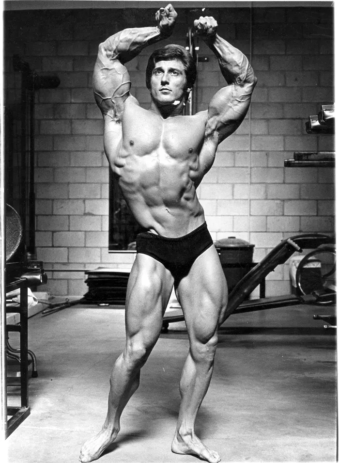 A dramatic low-angle double-biceps pose shot inside Gold's Gym, period equipment visible around him.