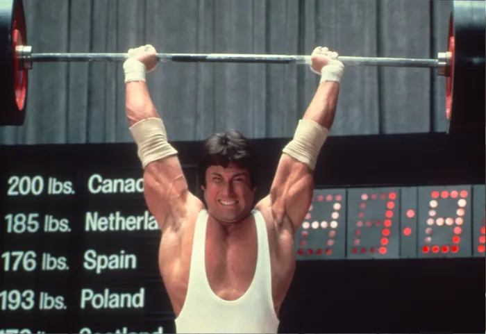 An overhead barbell lockout, gritted teeth, taped wrists, in front of an international weightlifting scoreboard.